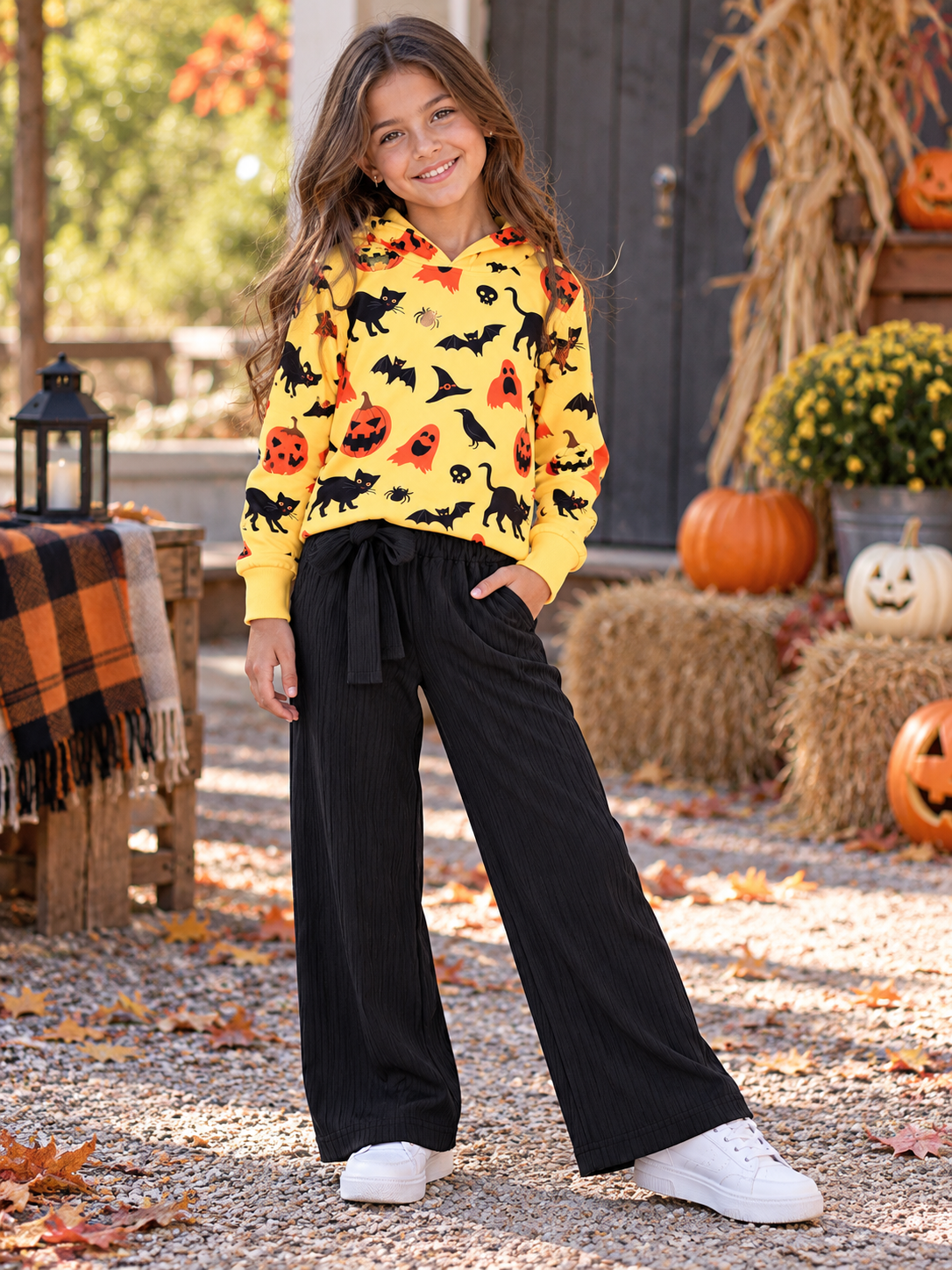 A young girl stands outdoors in a fall yard wearing a bright yellow long-sleeve hoodie covered in black bats, pumpkins, and ghosts, paired with black wide-leg pants and white sneakers.