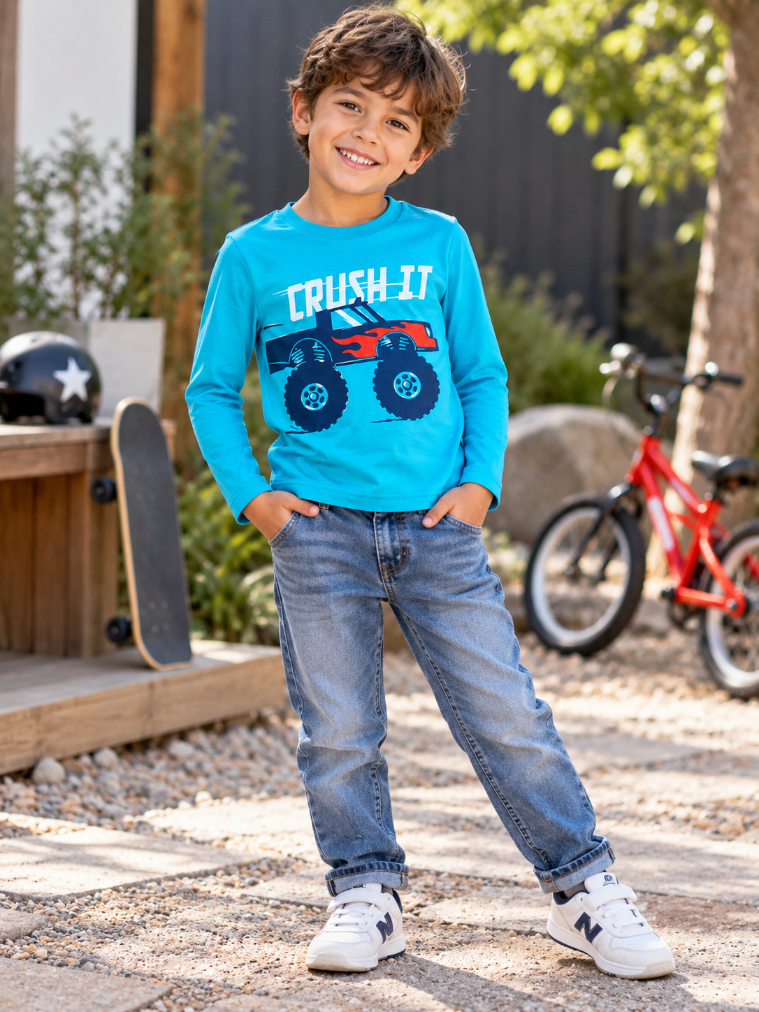 A young boy stands outdoors wearing a bright blue long-sleeve t-shirt with a red and blue monster truck graphic and the words 'CRUSH IT' across the front, paired with blue jeans and white sneakers.