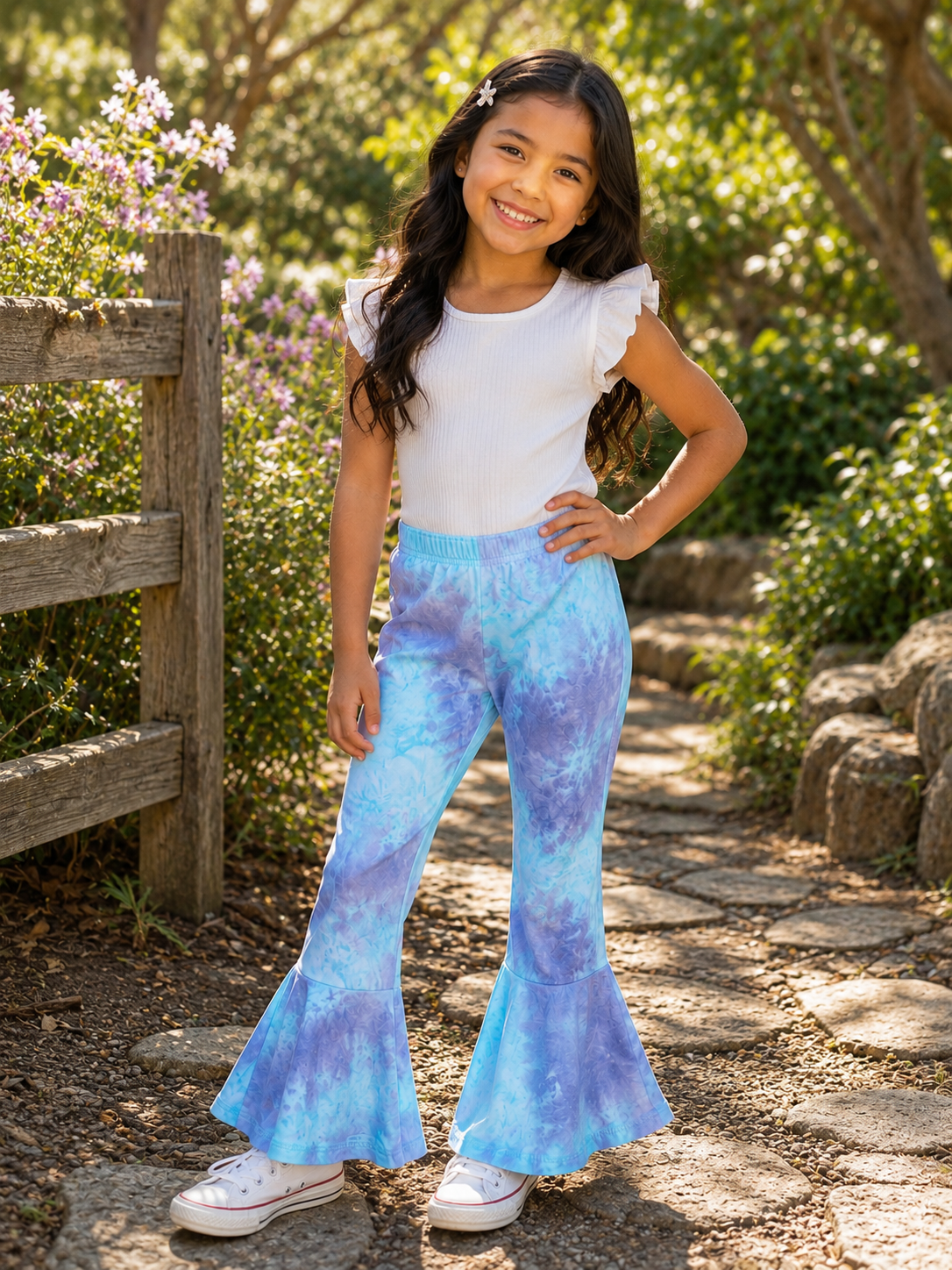 A young girl stands on a stone path in a garden, wearing blue and purple tie-dye flared pants, a white ruffle-sleeve top, and white sneakers.