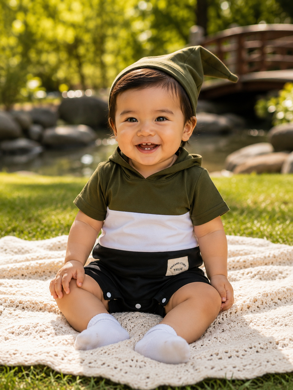 A smiling baby sitting outdoors on a cream crochet blanket, wearing an olive green and white color-block hoodie, black shorts, white socks, and an olive green pointed hat. The background shows a blurred green garden with a wooden bridge.
