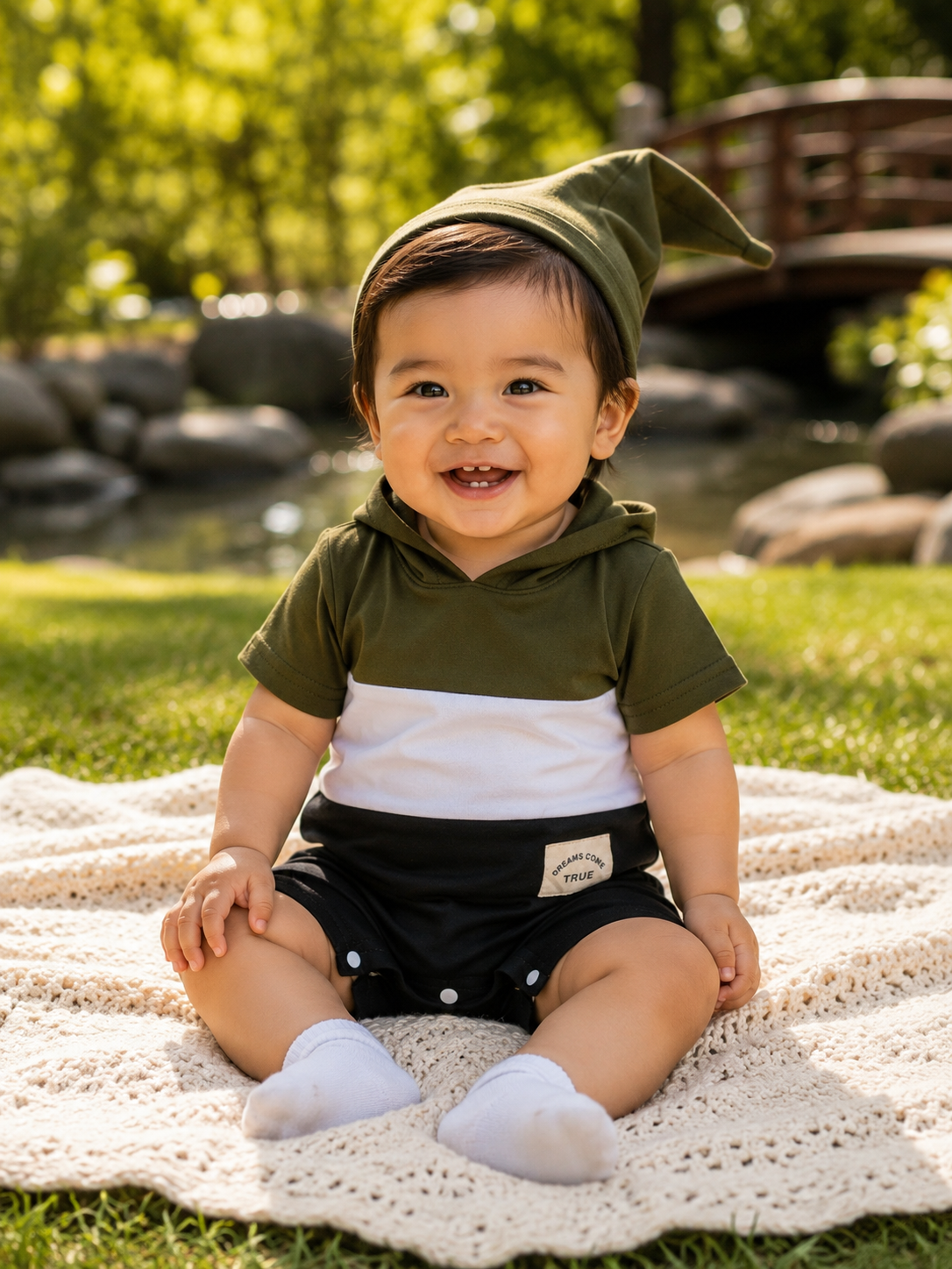 A smiling baby sitting outdoors on a cream crochet blanket, wearing an olive green and white color-block hoodie, black shorts, white socks, and an olive green pointed hat. The background shows a blurred green garden with a wooden bridge.