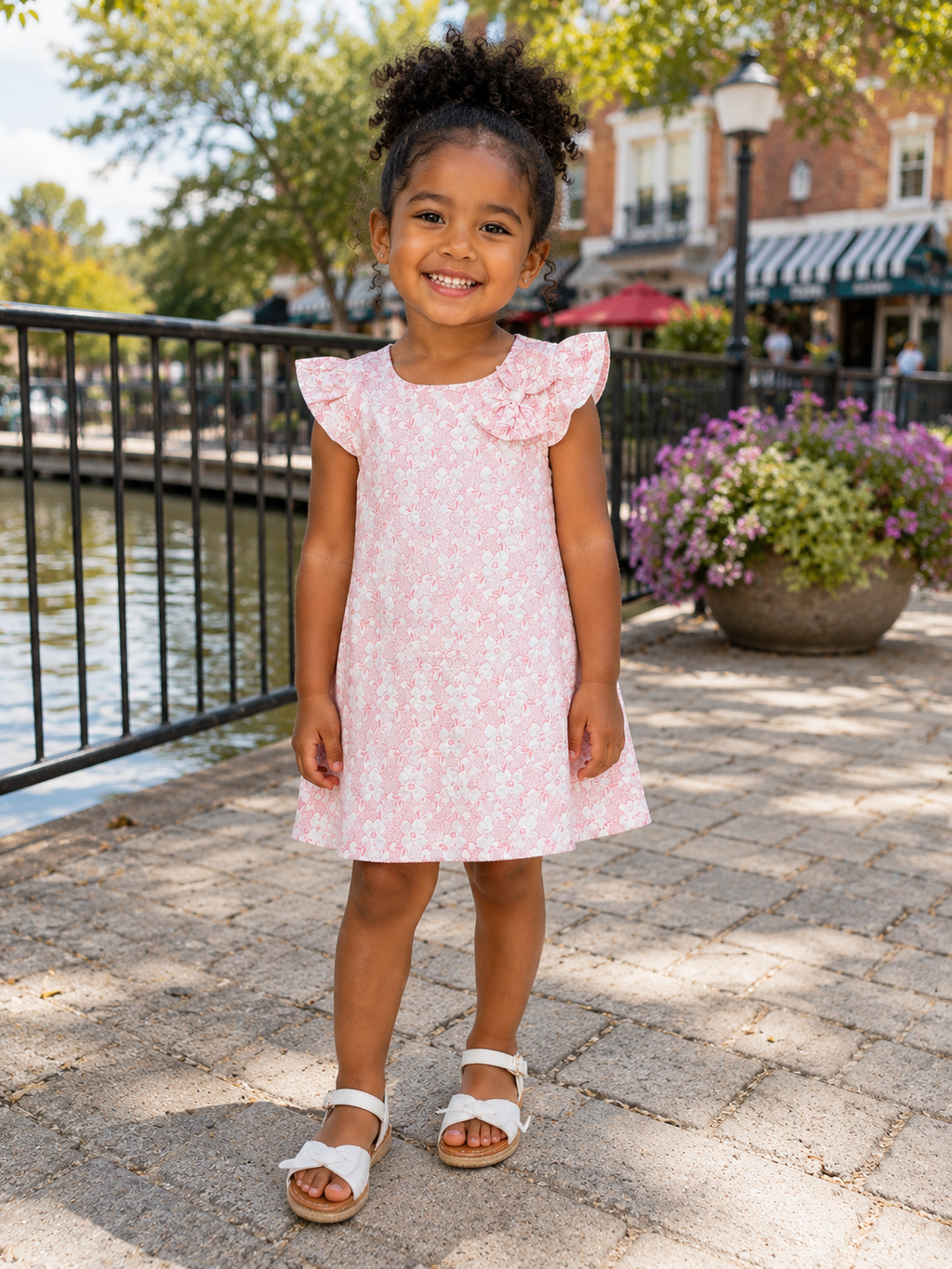 A young girl with curly hair stands on a stone walkway by a canal, wearing a light pink sleeveless dress with a white floral pattern and ruffled cap sleeves, paired with white sandals.