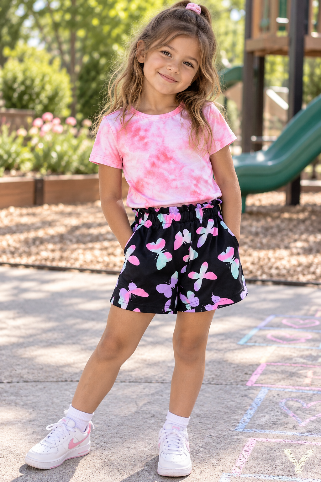 A young girl stands on a playground wearing a pink tie-dye t-shirt and black shorts with a pink, purple, and blue butterfly pattern. She is wearing white sneakers and standing in front of a green slide and playground equipment.