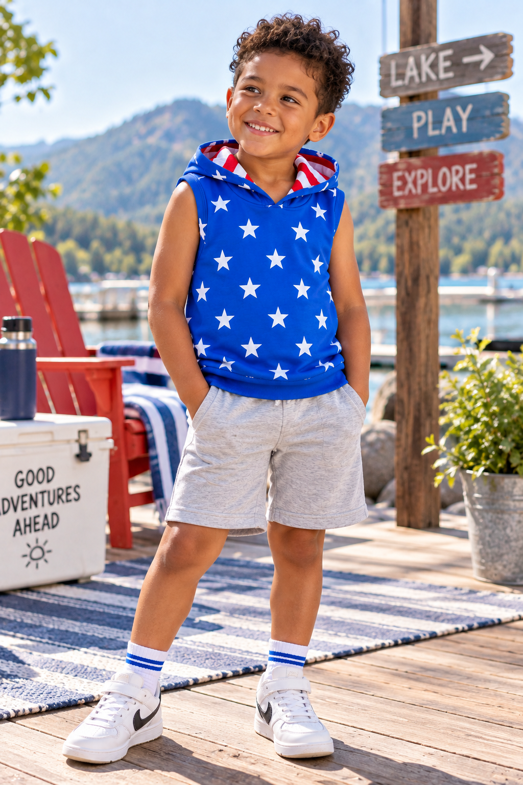 A young boy stands on a wooden dock by the water wearing a sleeveless blue hoodie with white stars and a red, white, and blue striped hood, paired with light gray shorts, white socks, and white sneakers.